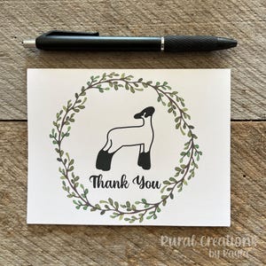 May include: A white greeting card with a black and white sheep illustration inside a green wreath. The words "Thank You" are below the sheep. A black pen rests above the card on a wooden surface.