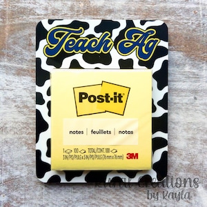 May include: A rectangular Post-it note holder with a black and white cow print border. The holder features the text "Teach Ag" in blue and yellow. A yellow Post-it note pad is in the center, with the Post-it logo and the words "notes, feuillets, notas".