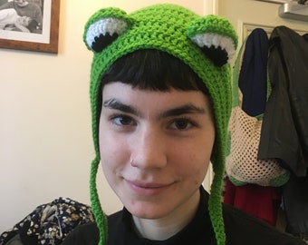 Könnte beinhalten: Ein grüner Häkelhut in Froschform mit weißen und schwarzen Augendetails. Der Hut hat lange grüne Bänder und ist handgefertigt. Ein lustiges und skurriles Accessoire.