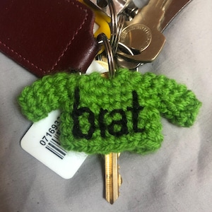 May include: A green crocheted sweater-shaped keyring with the word "brat" in black. The keyring is attached to a key and other keys, a maroon leather tag, and a yellow tag.