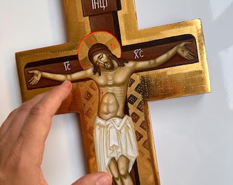 May include: A gold-colored wooden cross with a painted depiction of Jesus Christ. The cross features a detailed image of Christ with a halo, wearing a white cloth. The cross has text at the top and sides.