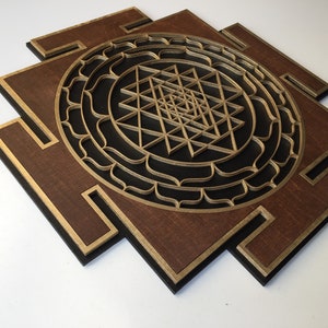 May include: A layered wooden wall art piece featuring a geometric design. The artwork has a dark brown base with gold and black accents. The central design is a complex, layered geometric pattern in gold.