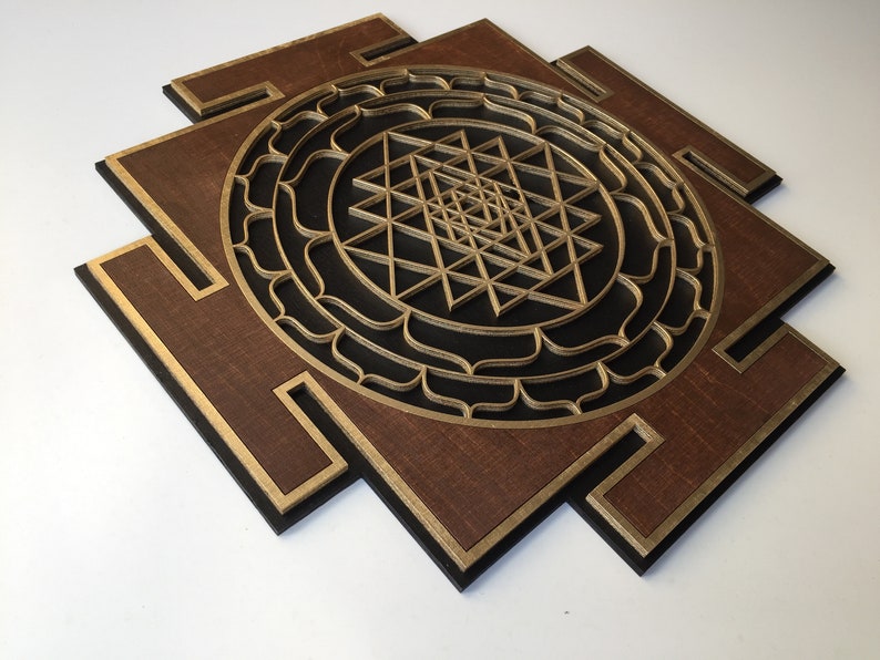 May include: A layered wooden wall art piece featuring a geometric design. The artwork has a dark brown base with gold and black accents. The central design is a complex, layered geometric pattern in gold.