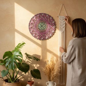 K&ouml;nnte beinhalten: Eine kreisf&ouml;rmige Mandala-Wanddekoration in Rosa-, Gr&uuml;n- und Braunt&ouml;nen h&auml;ngt an einer beigen Wand. Ein Makramee-Wandbehang und eine Frau sind ebenfalls zu sehen. Eine Monstera-Pflanze in einem geflochtenen Korb und Vasen mit getrockneten Pflanzen stehen auf einem Holzregal.