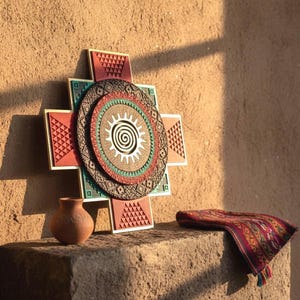 May include: A decorative wall hanging with a cross-like design, featuring geometric patterns in red, teal, and brown. A small brown vase and a patterned textile are on a stone surface. The wall is a warm, earthy tone.
