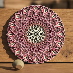 K&ouml;nnte beinhalten: Ein mehrschichtiges Holzmandala in Rosa, Rot, Schwarz und Wei&szlig;. Das kreisf&ouml;rmige Design weist komplizierte Blumenmuster auf und wird auf einer Holzoberfl&auml;che pr&auml;sentiert. Eine kleine Holzkugel befindet sich in der N&auml;he.