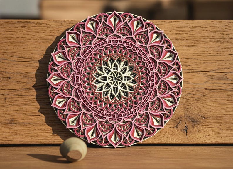 K&ouml;nnte beinhalten: Ein mehrschichtiges Holzmandala in Rosa, Rot, Schwarz und Wei&szlig;. Das kreisf&ouml;rmige Design weist komplizierte Blumenmuster auf und wird auf einer Holzoberfl&auml;che pr&auml;sentiert. Eine kleine Holzkugel befindet sich in der N&auml;he.