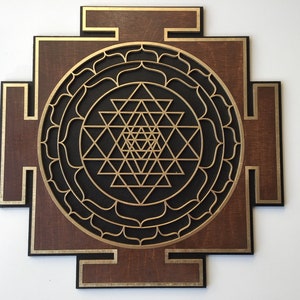 May include: A wooden wall art piece featuring a geometric Sri Yantra design. The artwork has a dark brown background with gold and black accents. The central design is a complex arrangement of triangles and circles.