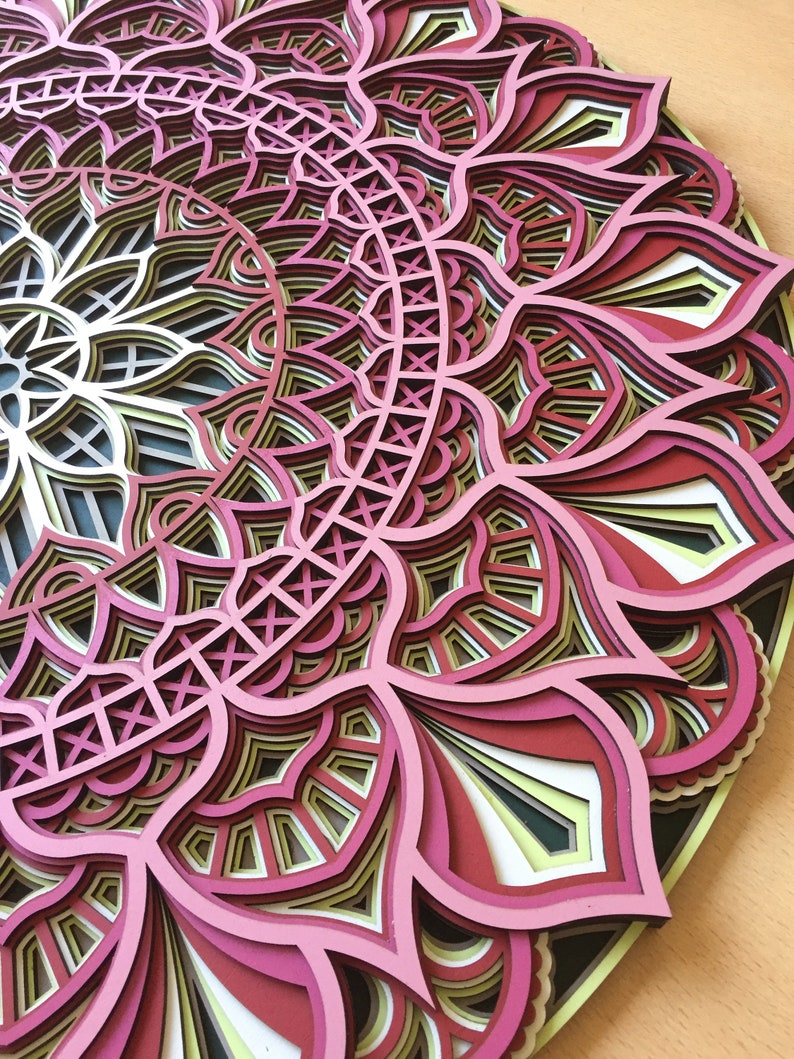 K&ouml;nnte beinhalten: Ein mehrschichtiges Mandala-Design in Rosa, Rot, Gelb und Wei&szlig;. Das kreisf&ouml;rmige Kunstwerk zeigt ein komplexes Muster aus floralen und geometrischen Formen, das einen 3D-Effekt erzeugt. Das St&uuml;ck wird auf einer hellen Oberfl&auml;che pr&auml;sentiert.