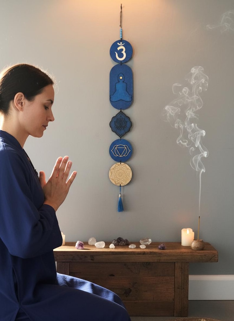 May include: A blue and gold chakra wall hanging with a tassel. The wall hanging features symbols and designs associated with meditation and spirituality. A woman is meditating in front of the wall hanging.