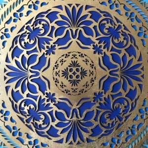 Könnte beinhalten: Eine runde Wanddekoration aus goldfarbenem Metall mit einem blauen und türkisfarbenen Design. Das Design zeigt komplizierte Ausschnitte von Blumen und Blättern.