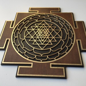 May include: A wooden wall art piece featuring a layered geometric design. The artwork has a dark brown base with gold and black accents. The central design is a gold-colored Sri Yantra symbol.