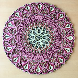 K&ouml;nnte beinhalten: Ein mehrschichtiges, kreisf&ouml;rmiges Mandala-Kunstwerk. Das Design weist komplizierte Muster in Rosa-, Schwarz-, Wei&szlig;- und Braunt&ouml;nen auf. Das St&uuml;ck wird auf einer hellen Holzoberfl&auml;che pr&auml;sentiert, die die detaillierte Handwerkskunst hervorhebt.