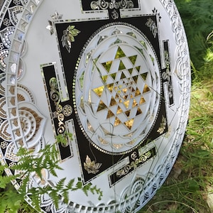 Op de afbeelding: Een witte en zwarte cirkelvormige mandala met een gouden Sri Yantra-ontwerp in het midden. De mandala is gemaakt van spiegeltegels en heeft een decoratieve rand.