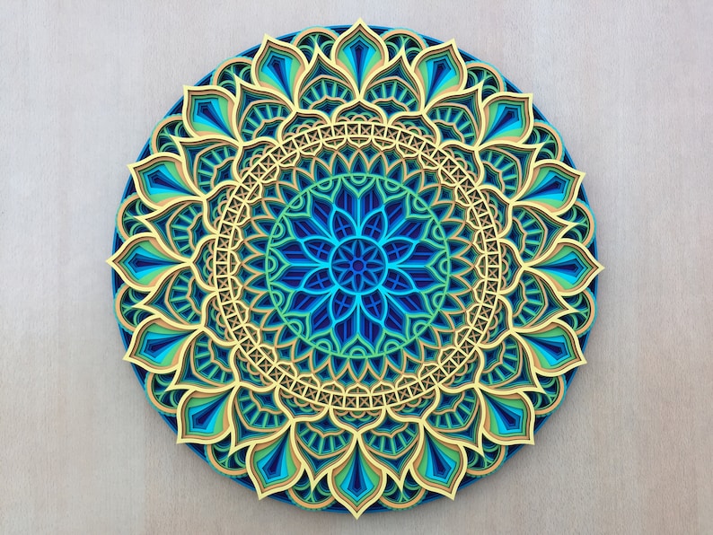 Op de afbeelding: Een blauwe, groene en gouden mandala met ingewikkelde patronen en lagen. De mandala is gemaakt van papier en heeft een 3D-effect.