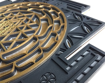 May include: A black and gold geometric wall art piece with a layered design. The piece features a circular mandala with a geometric pattern in the center, surrounded by a series of concentric circles.
