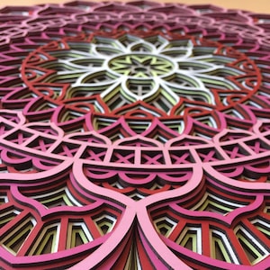 K&ouml;nnte beinhalten: Ein mehrschichtiges Holzmandala in Rosa-, Rot-, Wei&szlig;- und Gelbt&ouml;nen. Das komplizierte Design weist ein symmetrisches Blumenmuster mit mehreren Schichten auf, wodurch ein 3D-Effekt entsteht. Dieses dekorative St&uuml;ck ist ideal f&uuml;r Wandkunst.