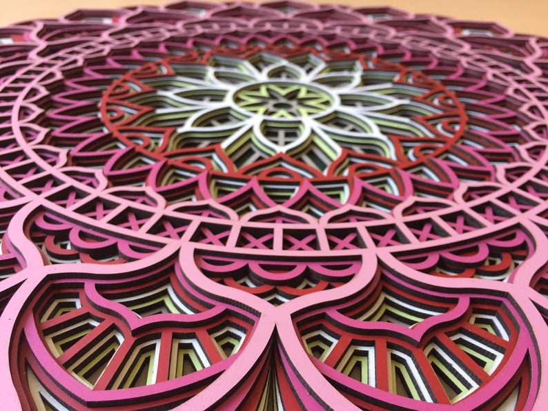 K&ouml;nnte beinhalten: Ein mehrschichtiges Holzmandala in Rosa-, Rot-, Wei&szlig;- und Gelbt&ouml;nen. Das komplizierte Design weist ein symmetrisches Blumenmuster mit mehreren Schichten auf, wodurch ein 3D-Effekt entsteht. Dieses dekorative St&uuml;ck ist ideal f&uuml;r Wandkunst.