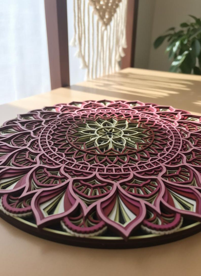 K&ouml;nnte beinhalten: Mehrschichtiges Holzmandala in Burgunder-, Rosa- und Cremet&ouml;nen. Das komplizierte kreisf&ouml;rmige Design weist detaillierte Ausschnitte auf. Das Mandala wird auf einer hellbraunen Oberfl&auml;che pr&auml;sentiert und zeigt seine Tiefe und Handwerkskunst.