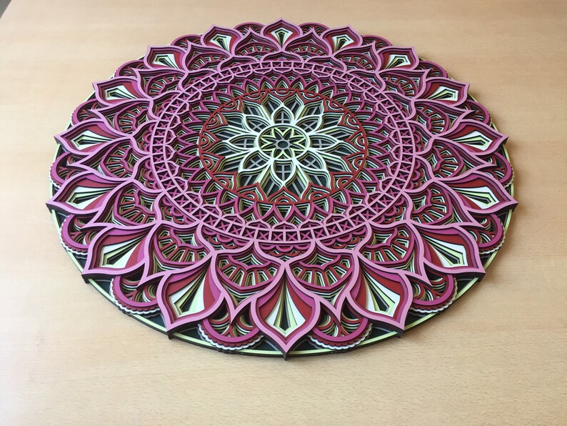 K&ouml;nnte beinhalten: Ein mehrschichtiges, kreisf&ouml;rmiges Mandala-Kunstwerk. Das Design weist komplizierte Muster in Rosa-, Rot-, Schwarz-, Gelb- und Wei&szlig;t&ouml;nen auf. Das Kunstwerk wird auf einer hellbraunen Oberfl&auml;che pr&auml;sentiert, die die detaillierte Handwerkskunst hervorhebt.