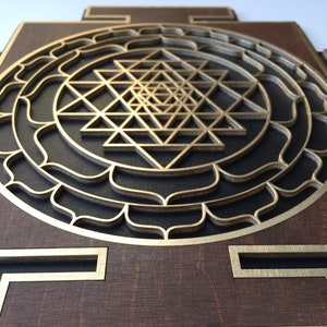 May include: A layered wooden wall art piece featuring a gold-colored Sri Yantra design. The artwork has a dark brown base with gold accents and a geometric pattern. The design is a symbol of spiritual significance.