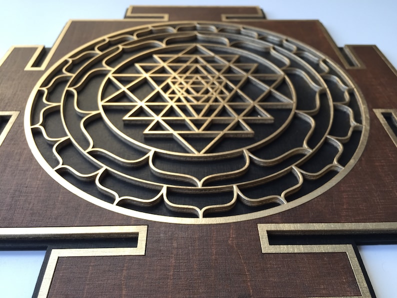 May include: A layered wooden wall art piece featuring a gold-colored Sri Yantra design. The artwork has a dark brown base with gold accents and a geometric pattern. The design is a symbol of spiritual significance.