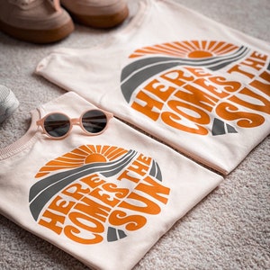 Op de afbeelding: Twee lichtroze T-shirts met de tekst "Here Comes The Sun" in oranje en grijs. De shirts zijn gevouwen en liggen op een lichtbruin tapijt. Een paar lichtbruine sneakers en een roze zonnebril zijn ook op de foto.