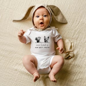 May include: A white baby onesie with a black and white illustration of two dogs and the text "Coco & Milo's New Best Friend". The baby is wearing a brown bunny hat.