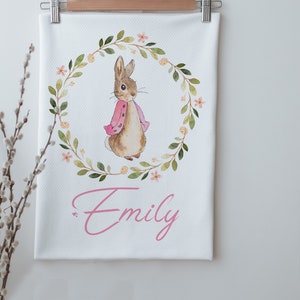 Peut inclure: Une serviette de cuisine blanche avec une illustration aquarelle d'un lapin brun portant une veste rose. Le lapin est entouré d'une couronne de feuilles vertes avec des fleurs roses. Le nom "Emily" est écrit en cursive rose sous le lapin.