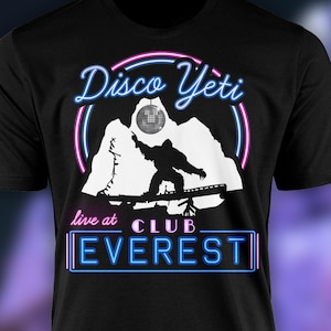 Op de afbeelding: Zwart T-shirt met een witte afbeelding van een yeti die snowboardt met een discobal erboven. De tekst "Disco Yeti" staat in roze en blauwe neonletters. De tekst "live at CLUB EVEREST" staat in blauwe neonletters.