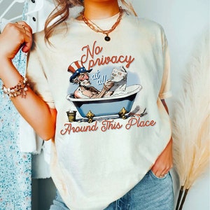 May include: A white t-shirt with a graphic of a cartoon Uncle Sam in a bathtub reading a newspaper. The text on the shirt reads "No privacy at all Around This Place".