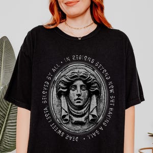 May include: Black t-shirt featuring a graphic print of a woman's face within a stone frame. The text surrounding the frame reads "BAIL ALL. DEAR SWEET LECT. IN REGIONS BEYOND NOW BUT HAVING."