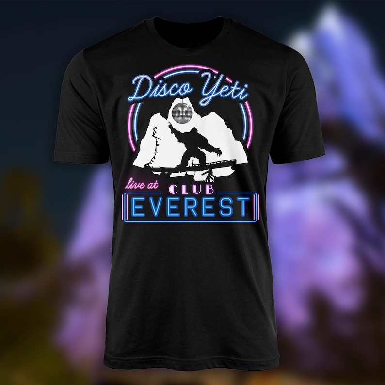 Op de afbeelding: Zwart T-shirt met een witte afbeelding van een yeti die snowboardt met een discobal erboven. De tekst "Disco Yeti" staat in roze en blauwe neonletters. De tekst "live at CLUB EVEREST" staat in blauwe neonletters.