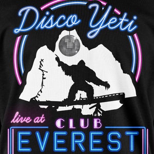 Op de afbeelding: Een zwart T-shirt met een witte afbeelding van een yeti die een discobal vasthoudt. De tekst "Disco Yeti" staat in een neonblauwe en roze lettertype boven de yeti. De tekst "live at CLUB EVEREST" staat in een neonblauw lettertype onder de yeti.