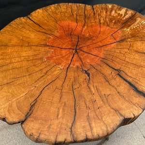 May include: A wooden table top with a natural wood finish and a reddish-brown center. The table top is round and has a rough, rustic texture.