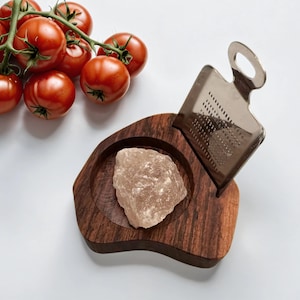 May include: A wooden salt cellar with a built-in grater for Himalayan salt. The cellar is made of dark wood and has a natural, organic shape. A large chunk of pink Himalayan salt sits in the center of the cellar.