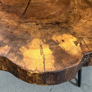 May include: A close-up of a wooden table top with a natural wood grain pattern. The table top is a live edge design with a dark brown stain. The table top is supported by three black metal legs.