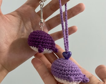 May include: Two purple crocheted whale keychains. One keychain has a silver spring ring and the other has a purple cord with a purple heart charm.