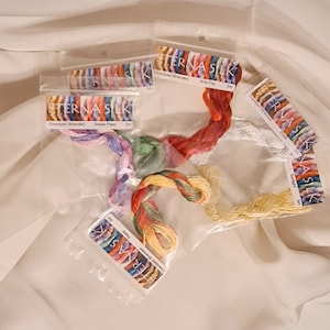 May include: Several packages of Eterna Silk overdyed stranded embroidery floss in various colors, including red, yellow, green, and purple. The floss is packaged in clear plastic and arranged on a white fabric surface.