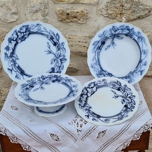 Puede incluir: Un juego de cuatro platos de cerámica blanca con un diseño floral azul. Los platos están colocados sobre un mantel de encaje blanco. El juego incluye dos platos grandes, un plato mediano y un plato pequeño sobre un pedestal.