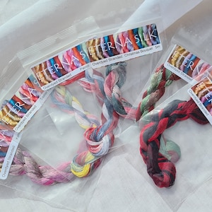 May include: Several skeins of colorful embroidery floss in clear plastic packaging. The floss is in various shades of pink, blue, green, and red. The packaging has the text "ETERNA SILK" and "Overdyed Stranded".
