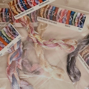 May include: Close-up of several skeins of overdyed stranded silk embroidery floss. The floss is in various colors, including pink, purple, and beige. The labels read "ETERNA SILK" and "Overdyed Stranded". Each skein is 5m in length.