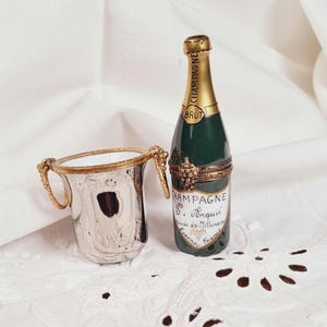 May include: A silver champagne bucket with gold handles and trim sits next to a green and gold champagne bottle-shaped trinket box. The box has a label that reads "Champagne S. Norquie". Both items are on a white, textured surface.