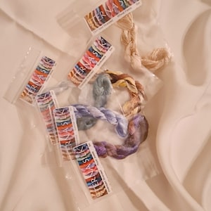 May include: Over-dyed stranded silk embroidery floss in various colors, packaged in clear plastic sleeves. The labels read "TERNA SILK" and include color names like "Wheat Fields" and "Summer Skies". The floss is arranged on a cream-colored fabric.