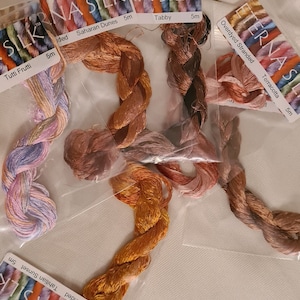 May include: Close-up of several skeins of silk embroidery floss in various colors. The floss is labeled with names like "Tutti Frutti," "Saharan Dunes," and "Tahitian Sunset." Each skein is 5 meters long and packaged in a clear plastic bag.