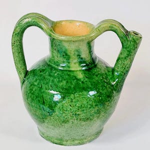 Può includere: Brocca in ceramica verde, piccola, con corpo arrotondato, due manici curvi e un beccuccio. La brocca ha una finitura lucida con variazioni di colore verde. L'interno è di colore giallo chiaro.