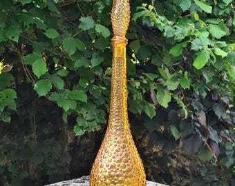 May include: A tall, amber-colored glass bottle with a textured, bubble-like surface and a pointed stopper. The bottle is displayed outdoors against a backdrop of green foliage, showcasing its vintage design.