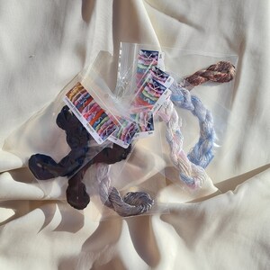 May include: Several skeins of colorful embroidery floss in clear plastic packaging. The floss colors include dark blue, gray, and pastel shades of pink, blue, and brown. Each package has a label with the brand name and color information.