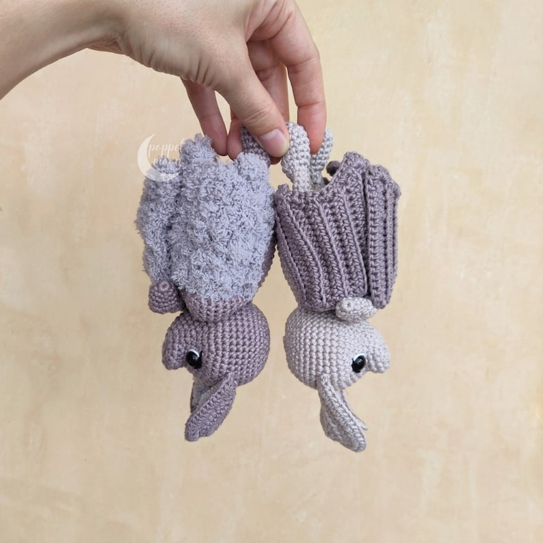 May include: Two crocheted bats hanging upside down. One bat is light purple and the other is a darker shade of purple.