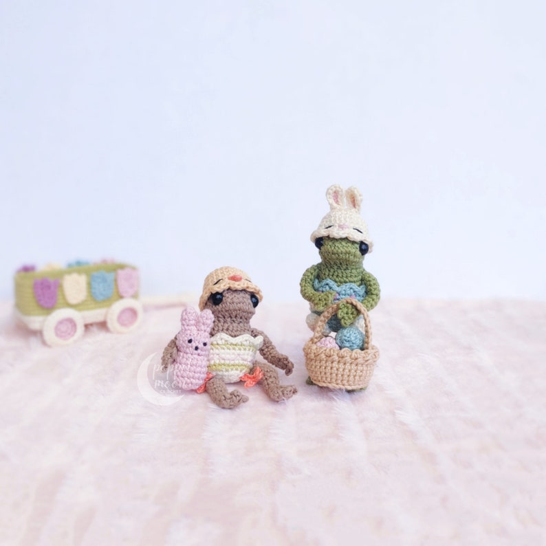 May include: Two crocheted Easter bunny figurines. One is green and holding a basket of colorful eggs. The other is brown and wearing a yellow hat, holding a pink bunny.